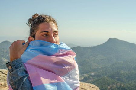 Picture of Close Up of Transgender Long Hair Man with Blue Eyes Covering his Face with LGTB Trans Flag Pink and Blue at the Mountain Cliffの写真素材