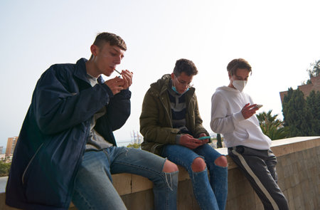 A closeup of three young Caucasian and Hispanic guys sitting outdoors smoking and using their phones wearing sanitary masksの写真素材