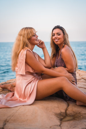 A couple of attractive smiling caucasian girls in summer dresses sitting on orange rocks against the background of the seaの写真素材