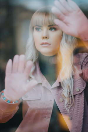 A young blonde caucasian female in a pink coat standing behind d a glass windowの写真素材