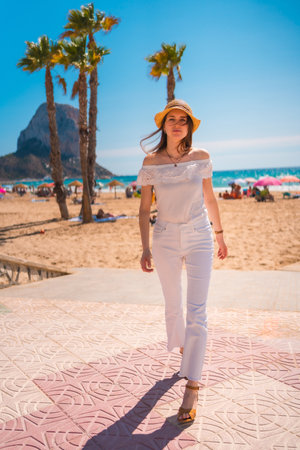An attractive young woman from Spain dressed in white with a straw hat walking along the beach of Calpe, Valencia, Spain.の写真素材