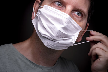 Close up of a caucasian man putting on a medical face mask with one hand stretching the elastic band and putting the protective mouthpiece onの写真素材
