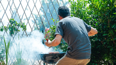 A back view of a person grilling the sausages on the charcoal grill in the backyardの写真素材
