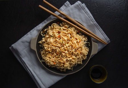 A top view of noodles with sesame seeds, teriyaki, chopsticks, and a  sauce on black background