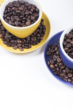 set of two ceramic cups with their plates one yellow and one blue filled with coffee beans placed diagonally on a white background, vertical photoの写真素材