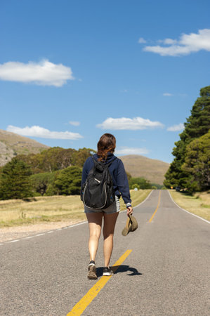 A vertical shot of a middle-aged Argentinian woman holding her cap and walking on the mountain road