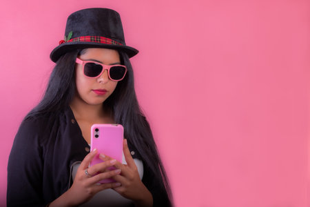 A beautiful Latin female in sunglasses and a hat chats with a friend by phone next to a pink wallの写真素材