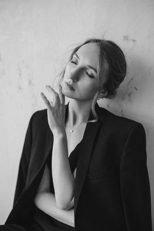 A grayscale shot of an attractive young European female in an elegant black suit posing at the camera with eyes closed and holding hand under chinの写真素材