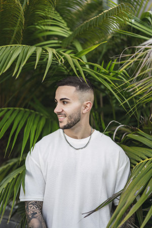 A vertical shot of a smiling European man in a white t-shirt posing against palm treesの写真素材