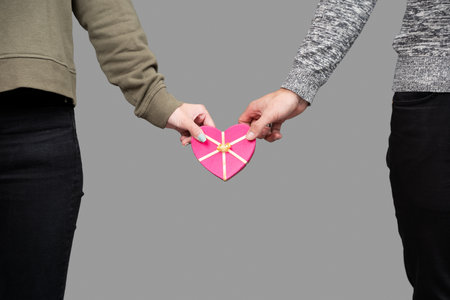Two people's hands holding a pink heart-shaped box on gray background
