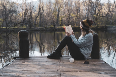 a young woman sits on a jetty above the lake, she is reading a bookの写真素材