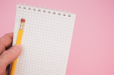 A closeup shot of a hand holding a pencil with a notebook on a pink backgroundの写真素材