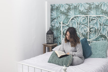 woman in bed reading a book, brunette woman in gray pajamas reads a book sitting on a bed with a...