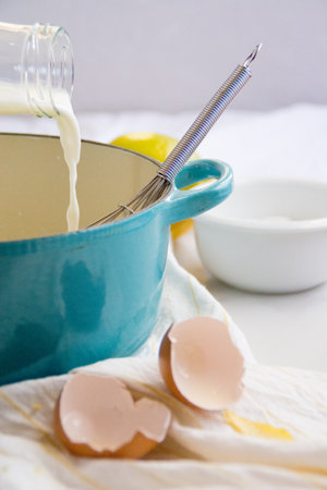 A vertical shot of milk pouring in a casserole with a whisk, and eggshells on white fabricの写真素材
