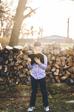 A little boy reading the holy bible while standing in front of stacked firewood in a fieldの写真素材