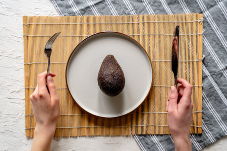 A top view of fresh whole avocado on a white plate with someone holding a fork and knife