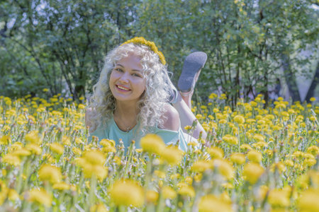 A beautiful and happy young girl in a green dress lies on a yellow lawn. There is a wreath of dandelions on her head. Looks into the camera.の写真素材