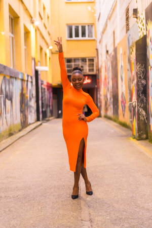 Fashionable urban style with a black African girl in an orange dress and black heels on a city street. Smiling and arm upの写真素材