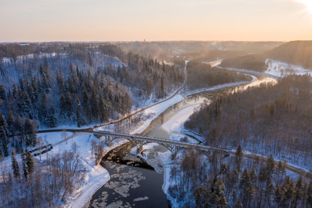 An aerial shot of a curvy road over a flowing river through the snow-covered forestの写真素材