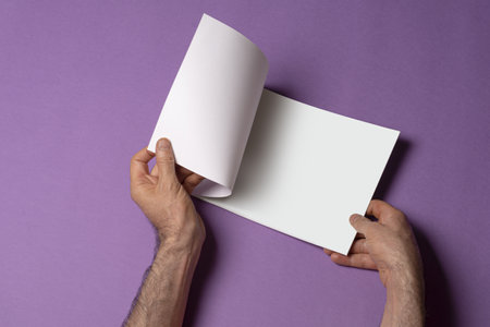 A closeup shot of a male hand holding a closed book-catalog with a blank cover on purple background