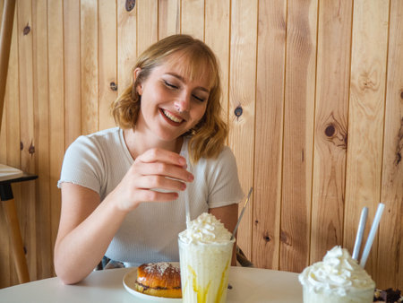 A beautiful blonde Caucasian woman from Spain sitting at a white table with two milkshakes and a donut in front of herの写真素材
