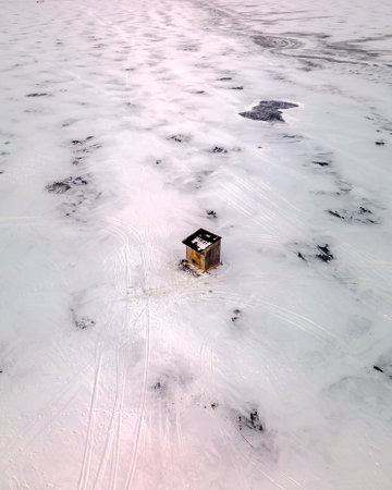 An aerial view of a wooden small hut in the middle of a frozen snow-covered field with ski tracksの写真素材