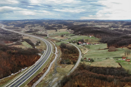 An aerial view of traffic roads amid agricultural fields and forests with residential buildings under a cloudy skyの写真素材