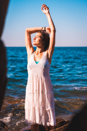 A young Caucasian woman enjoying looking at the sun with her eyes closed in a white dress and wet hair sitting on a rock in the sea, lifestyleの写真素材