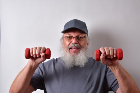 A happy American man with glasses and cap holding dumbbells and training on a white wall backgroundの写真素材