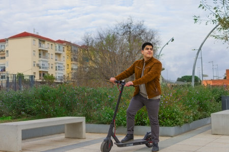 A young Spanish guy posing on an electric scooter on a square with benchesの写真素材