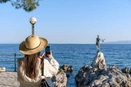 Young female tourist standing on rocky coastline with statue of a maiden with seagull in Opatija, Croatia.の写真素材