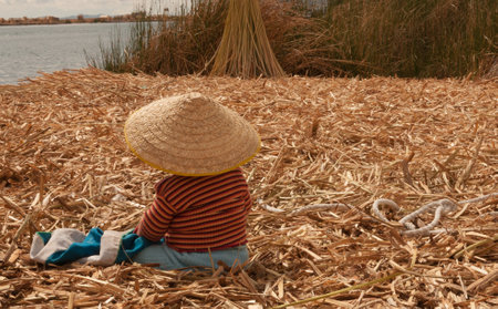 A child in a wicker conical hat sitting on the coast of the Uros Island, Peruの写真素材