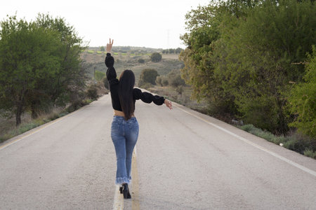 A back view of a young girl with long hair wearing blue jeans and a black cropped top sweater standing on an asphalt road making a victory gestureの写真素材