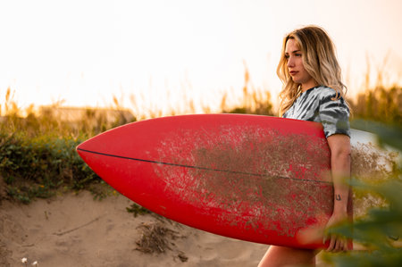 A young blonde female surfer walking with her surfing board at the beachの写真素材
