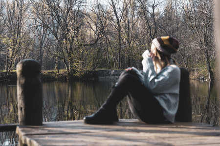 a selective focus shot of a woman relaxing in the wooden pier of the lake with a cup of coffeeの写真素材