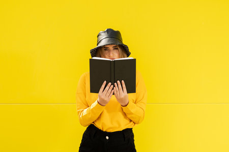 A closeup shot of a young Caucasian lady in yellow with a black hat holding an open notebookの写真素材