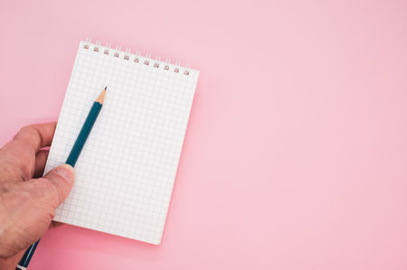 A closeup shot of a hand holding a pencil with a notebook on a pink backgroundの写真素材