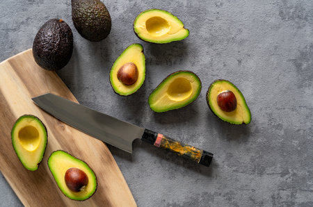 A top view of fresh halved and whole avocados on a grey surface, with a cutting board and knife