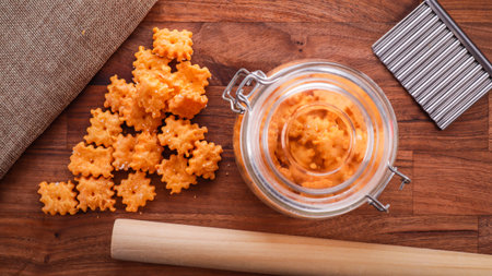 An overhead shot of bite size cheese crackers in a jar and on a wooden table with a rolling pin and...