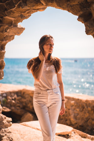 A young attractive woman from Spain dressed in white posing for the camera by the sea in a stone windowの写真素材
