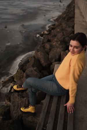 A closeup of a beautiful and young Ukrainian stylish female sitting on the wooden bench on the coastの写真素材