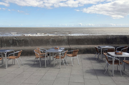 A cafeteria outside on pavement along the sea, displaying thatch chairs around small metal tables