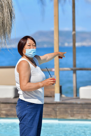 A vertical shot of an Asian female in a medical mask pointing at the seaの写真素材