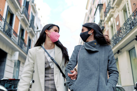 Two happy women talking and walking in the street with their face masks on.の写真素材