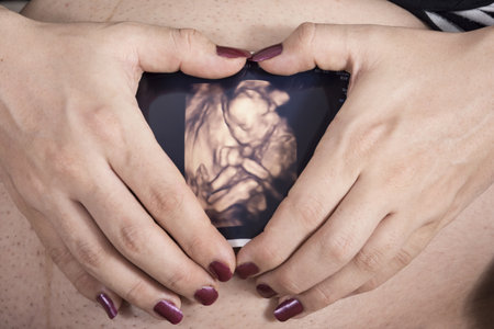 A mother holding baby sonography image in front of belly creating a heart-shape border with fingers