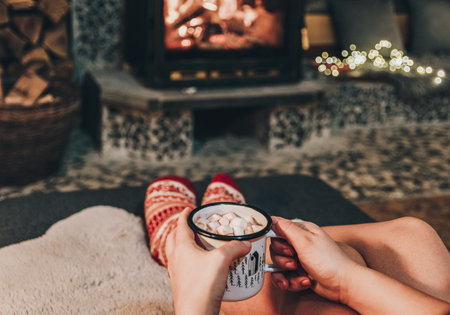 A closeup of a woman's feet in red woolen socks stretched out to the cozy fireplace holding a cup of coffee full of marshmallowsの写真素材