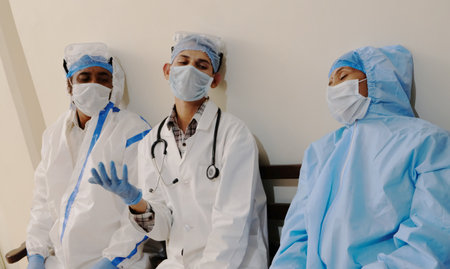 A group of tired South Asian doctors in full medical gear sitting on a hospital benchの写真素材