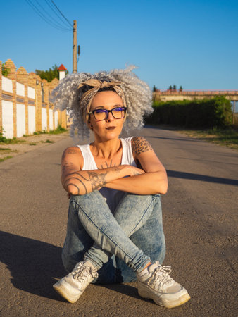 A shallow focus of a cool adult woman with white curly hair and tattoos sitting on the roadの写真素材