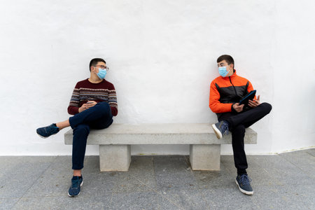 Stock photo of teenagers wearing face masks due to covid19 using tablet and talking while sitting in a bench.の写真素材