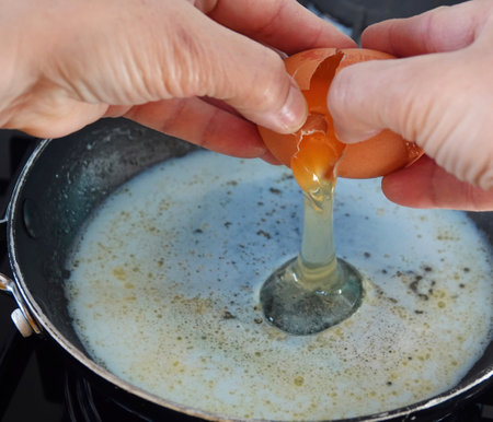 A closeup of an egg being cracked and poured into a pan on the oven - making scrambled eggs in the kitchenの写真素材